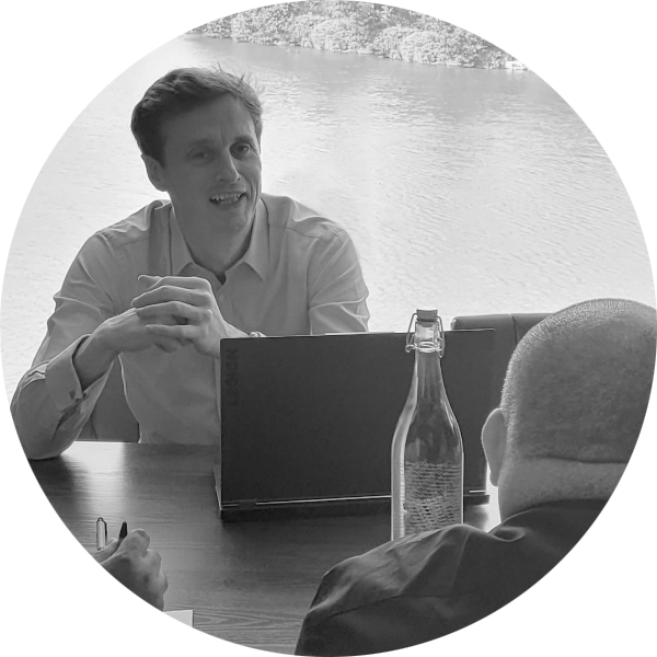 A smiling man at a laptop in a meeting with a second man. There is a view of a lake out of the window behind them.
