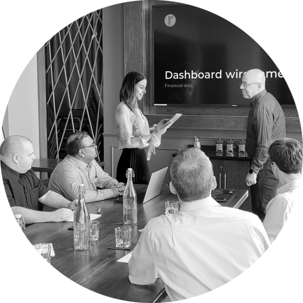 People sat around a table, watching a Powerpoint presentation of Dashboard Wireframes. A woman is standing with a tablet, talking to the man doing the presentation