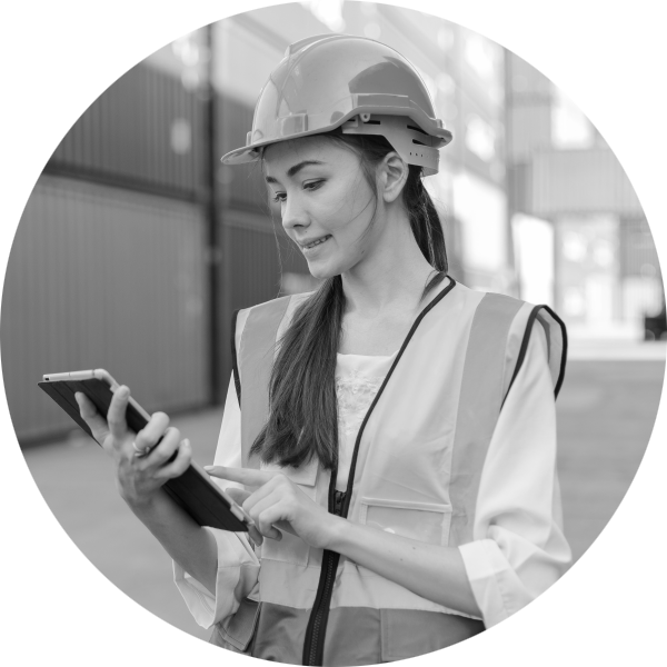 Woman looking at a tablet device, dressed in high visibility clothing with a hard hat in a storage area full of shipping containers